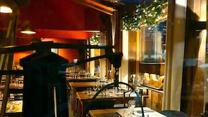An indoor dining area with wooden tables and chairs, a bar area with bottles and glasses, and a view through a window with greenery outside. - Chalet Hotel Du Bois (Les Houches)