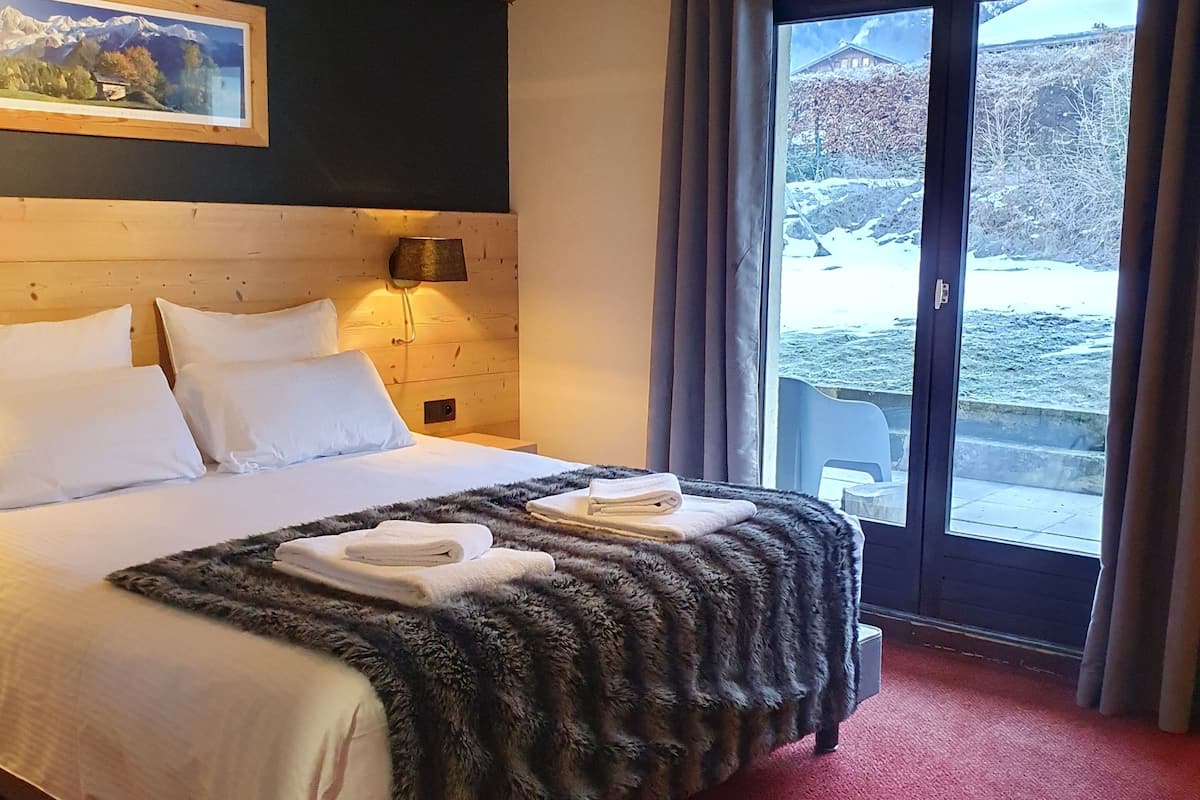 Une chambre d’hôtel avec un grand lit, une tête de lit en bois, un tableau au mur, une table de chevet et une vue sur un paysage enneigé par la fenêtre.