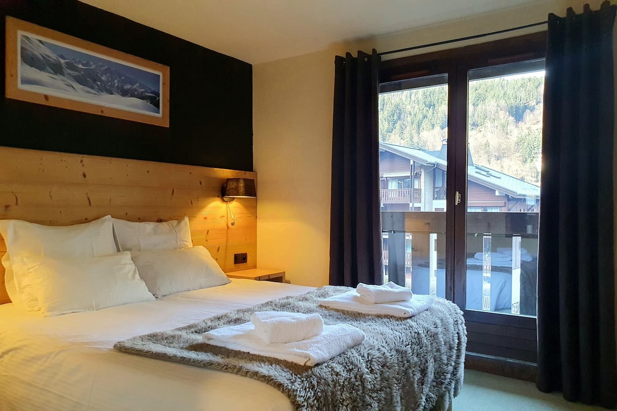 Un lit bien fait, avec du linge de lit blanc et une couverture moelleuse, une tête de lit en bois, une lampe de chevet et une fenêtre avec des rideaux.