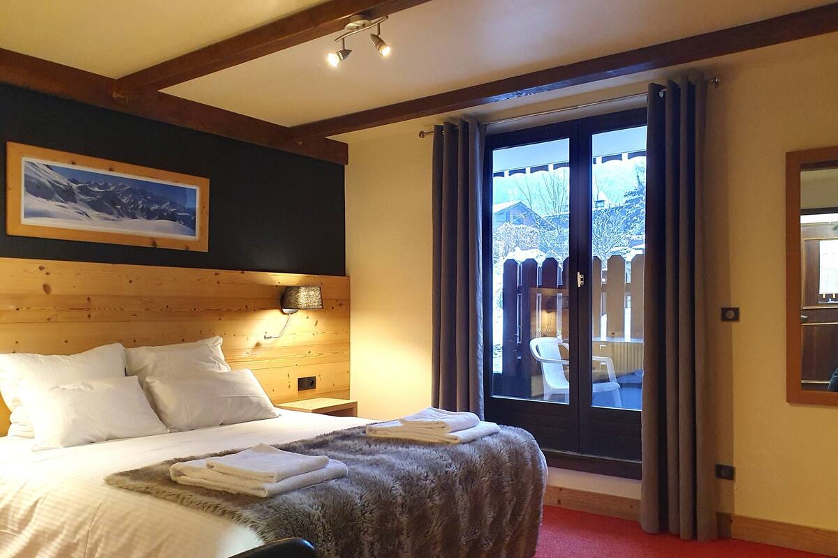Une chambre d’hôtel avec une tête de lit en bois, un grand lit avec du linge de lit blanc, une couverture grise moelleuse et une moquette rouge. Il y a une fenêtre donnant sur un paysage enneigé et un balcon avec une chaise.