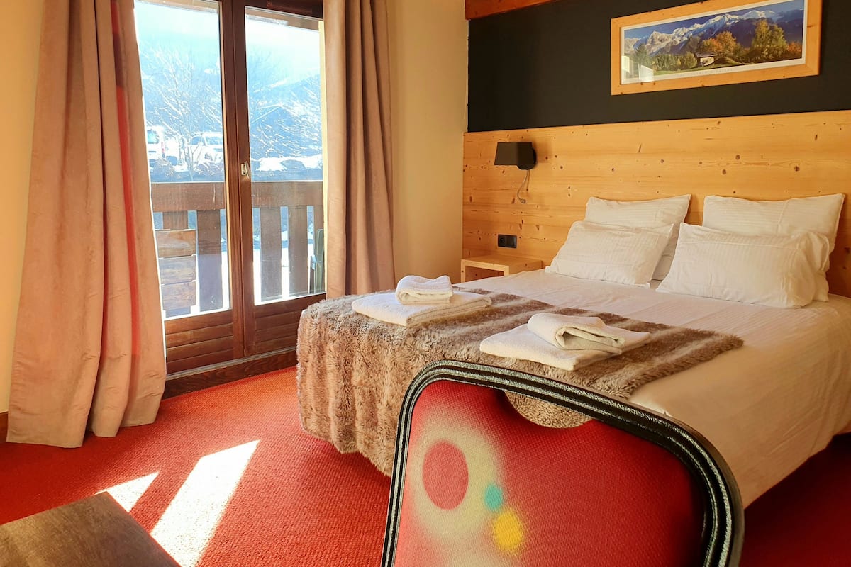 Une chambre d’hôtel avec une tête de lit en bois, un lit recouvert de draps blancs, une moquette rouge et une vue sur une montagne enneigée par une porte vitrée.