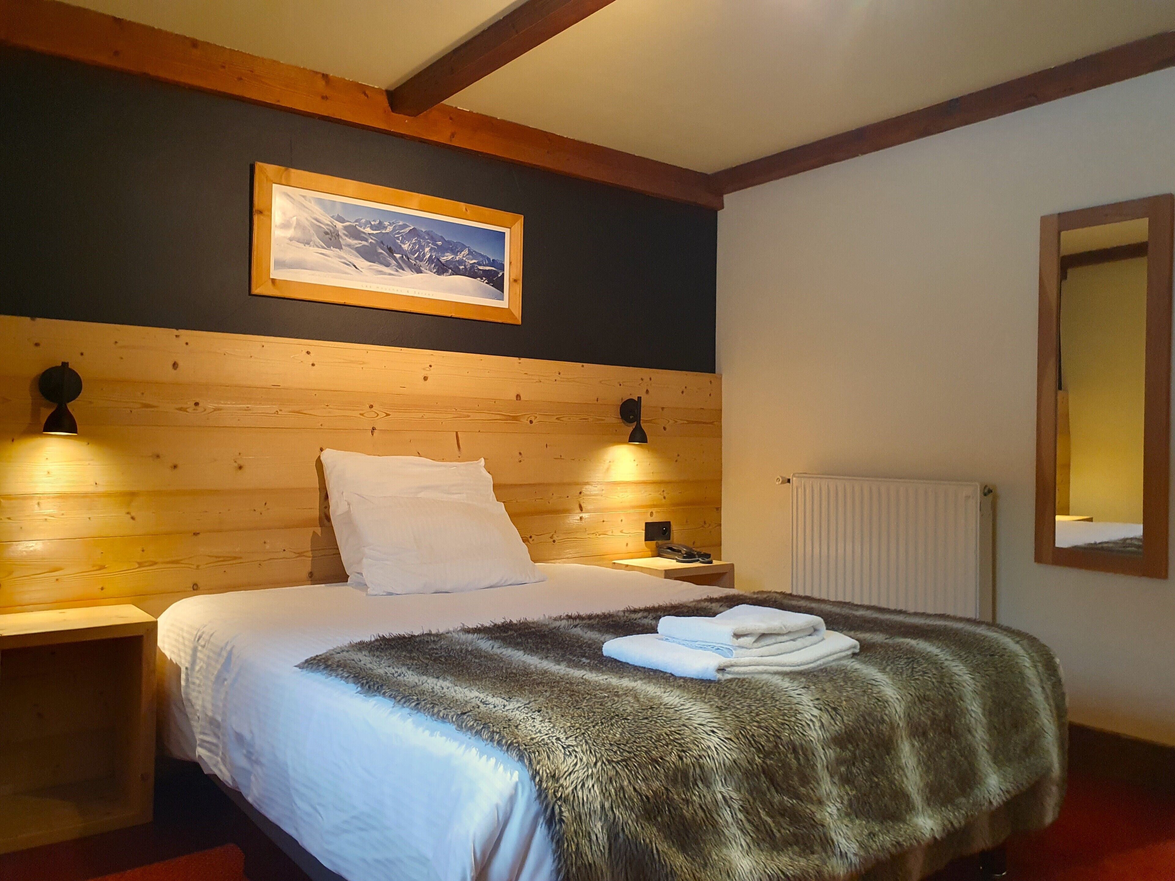 Una habitación de hotel con cabecera de madera, un cuadro enmarcado en la pared, dos lámparas de noche, una cama blanca con manta mullida y un radiador.