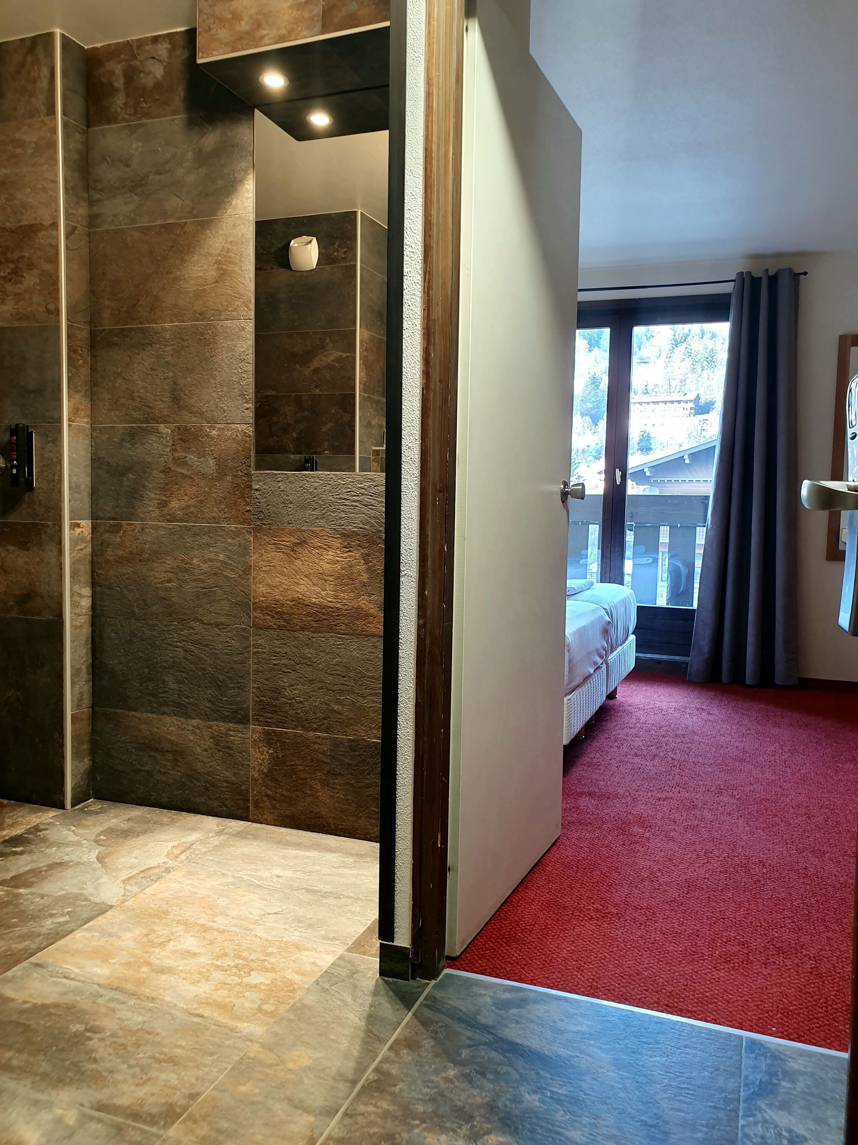 Habitación de hotel con alfombra roja, un ventanal con vista a un edificio y una puerta de madera que da a un balcón.