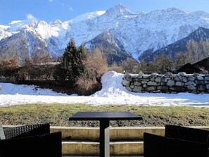 Une table et des chaises disposées dans un décor extérieur enneigé, avec un mur de pierre et une vue sur des montagnes recouvertes de neige.