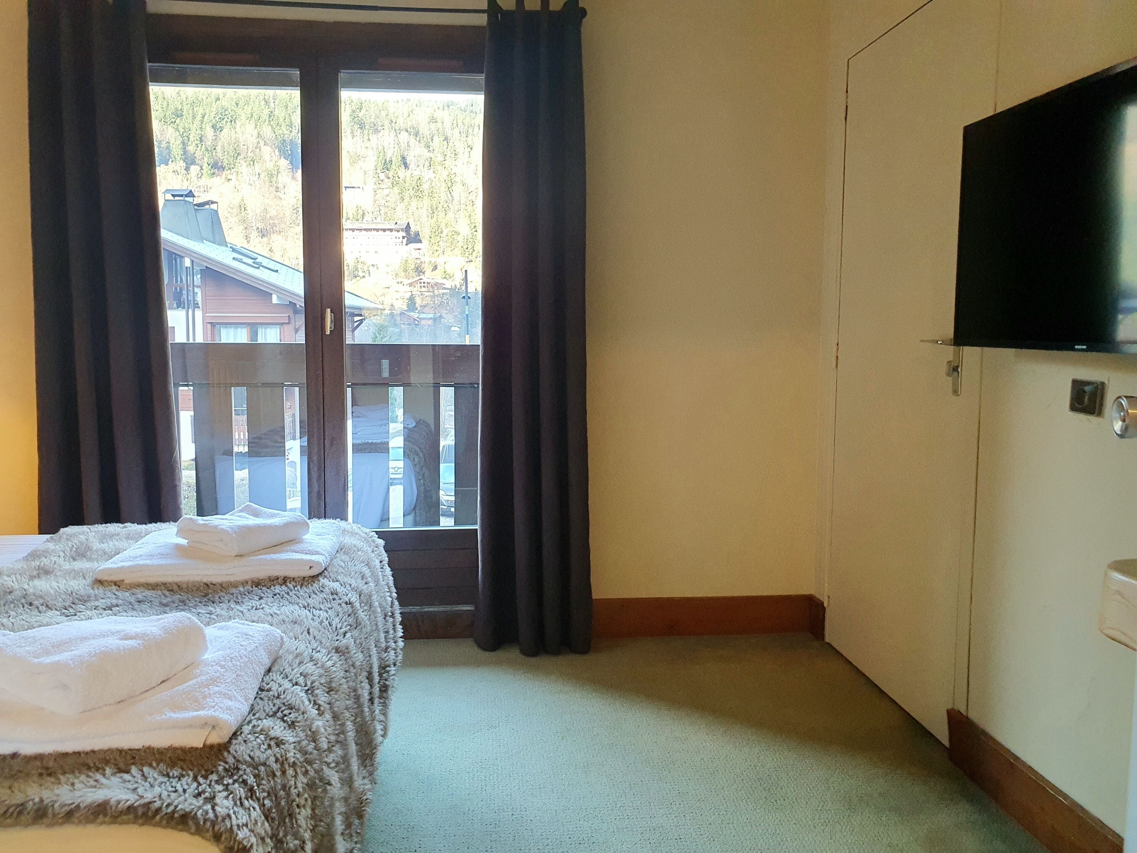 A hotel room with a bed, a TV, and a view of buildings through a window.