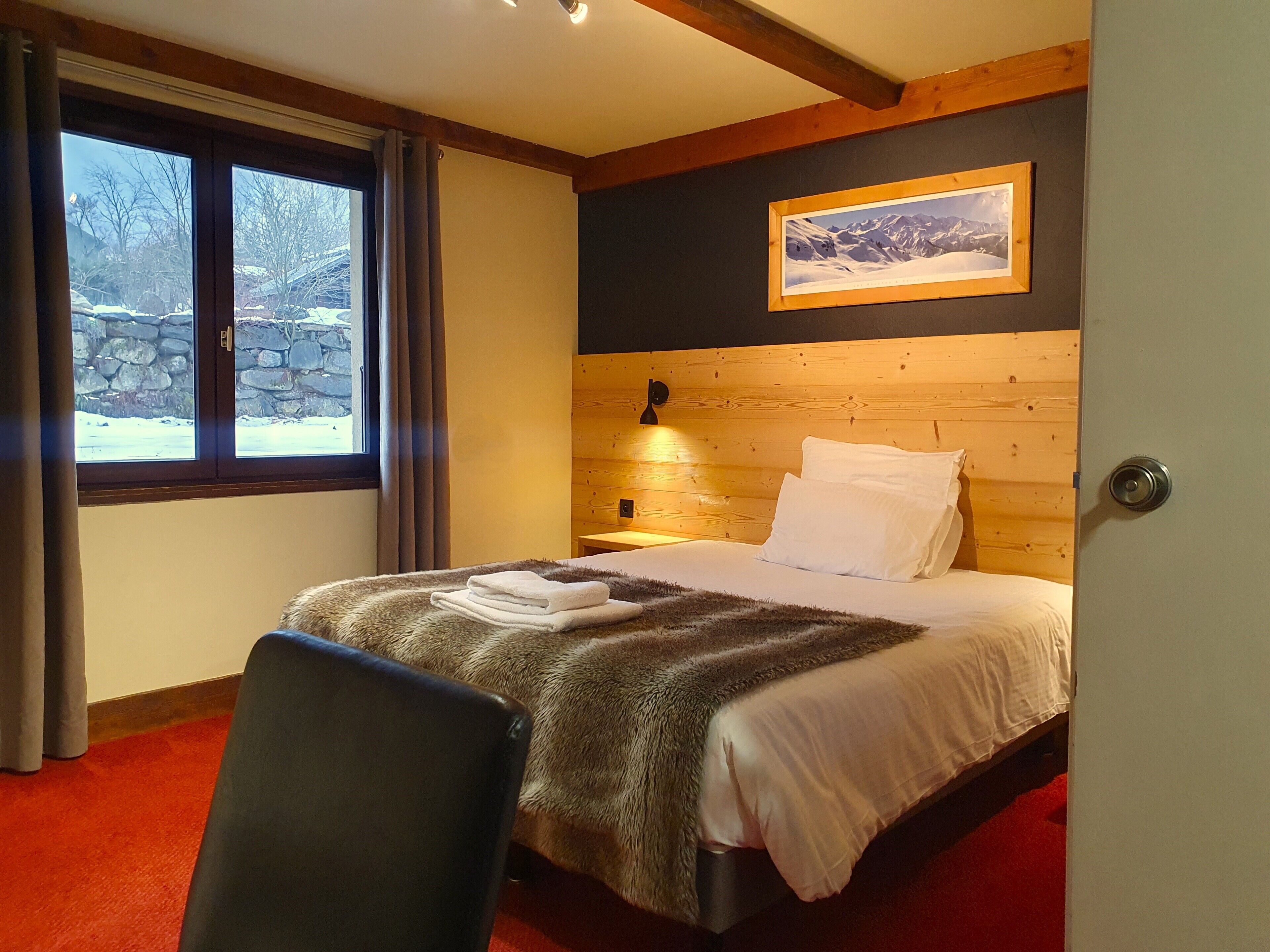 Ein Hotelzimmer mit einem hölzernen Kopfteil, einem Bett mit weißen Laken, einem roten Teppich, einem Sessel und einem Fenster mit Blick auf eine verschneite Landschaft.