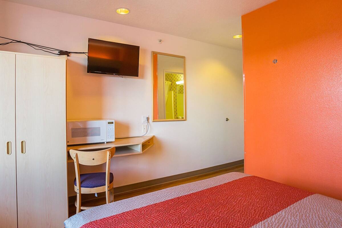 A hotel room with a bed, a desk with a microwave, a chair, a TV mounted on the wall, and a mirror.