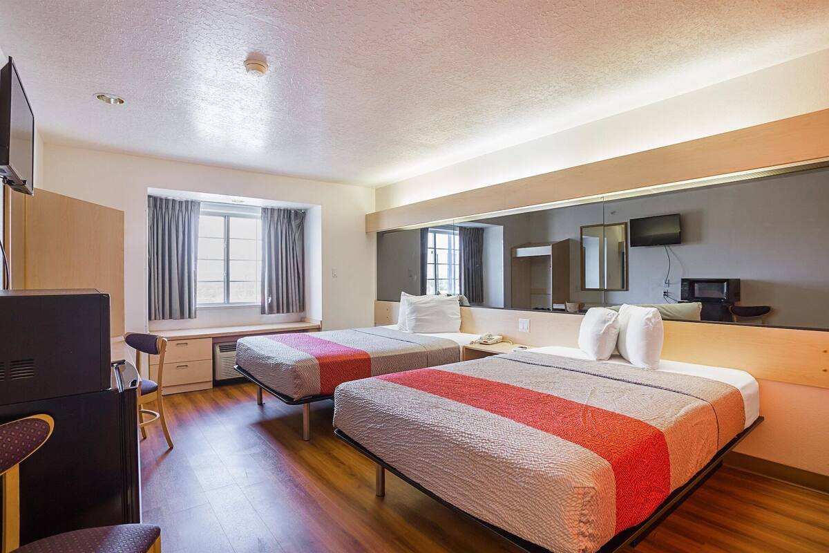A hotel room with two beds, a desk, a chair, a TV, a mirror, and a window with curtains.