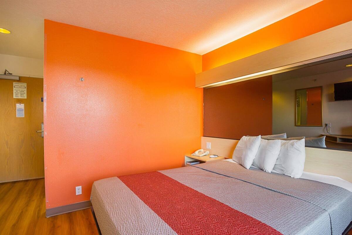 A hotel room with an orange wall, a bed with a patterned blanket, a bedside table with a phone, and a mirror on the wall.