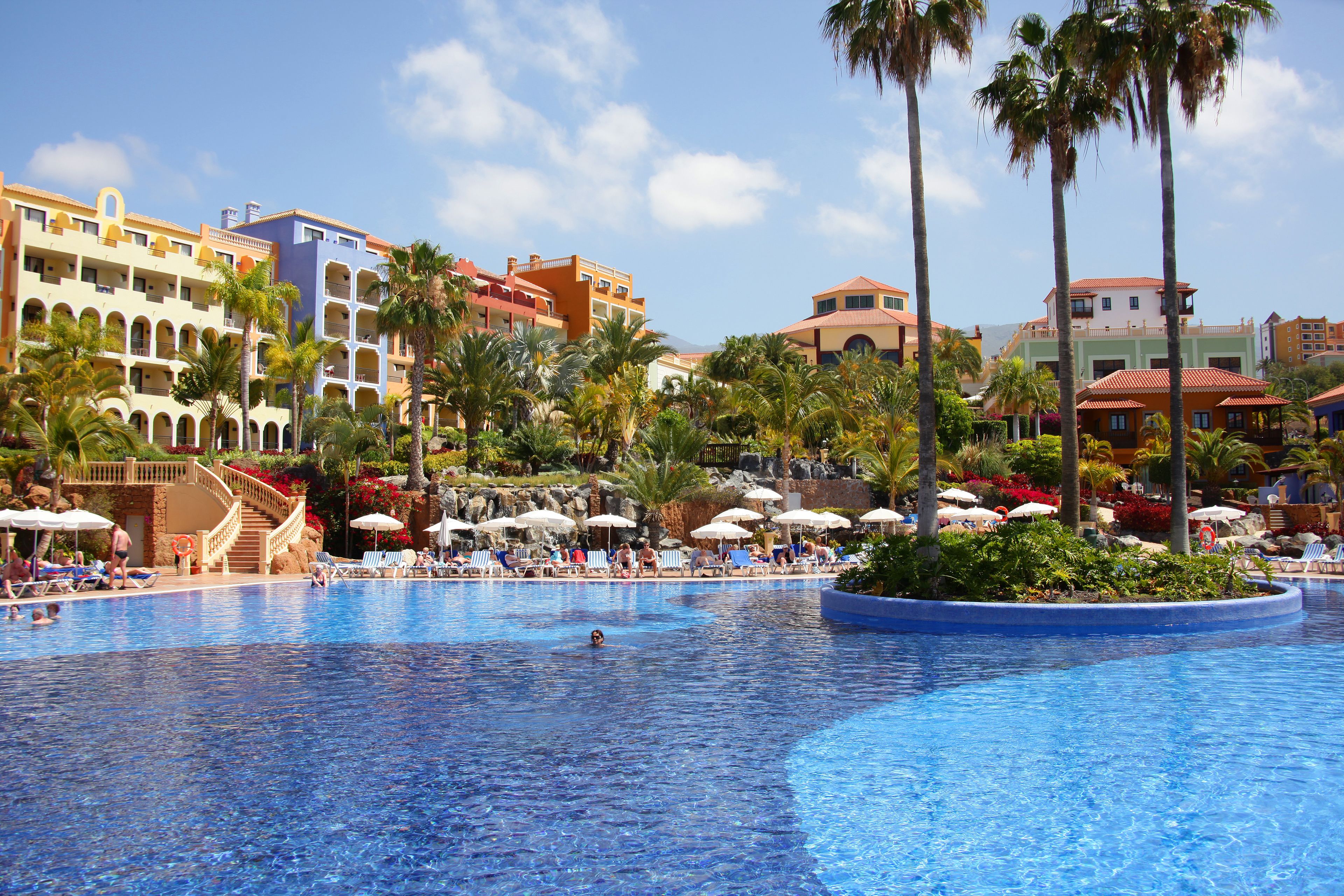 Una amplia piscina con sillones de playa y sombrillas, rodeada de palmeras y edificios de colores.