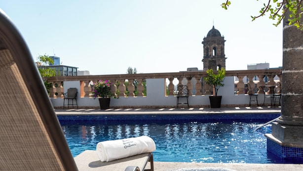 A rooftop pool with a view of a historic tower, lounge chairs, and potted plants.