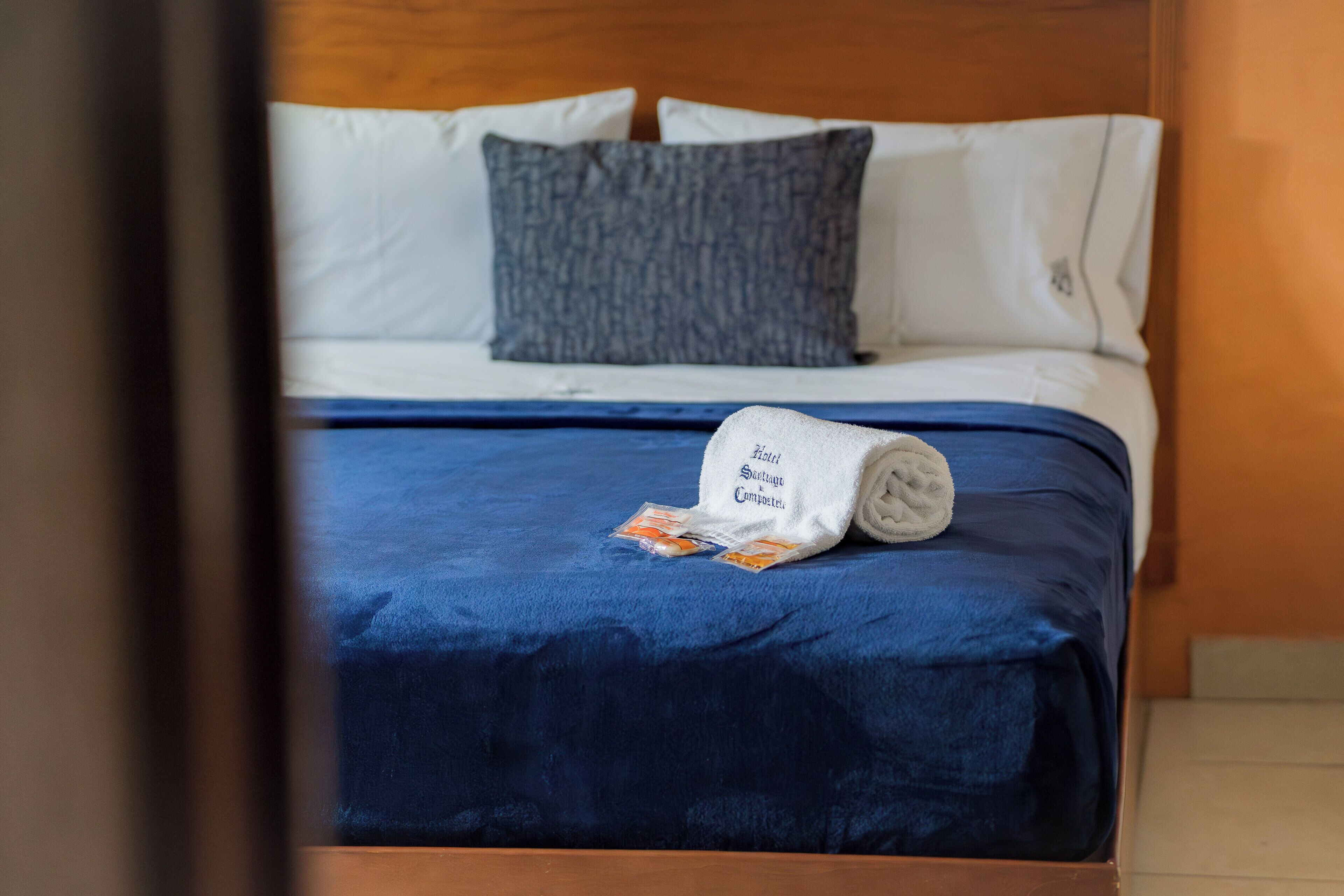Un lit bien fait, avec une couverture bleue et une serviette enroulée sur laquelle on peut lire un texte.