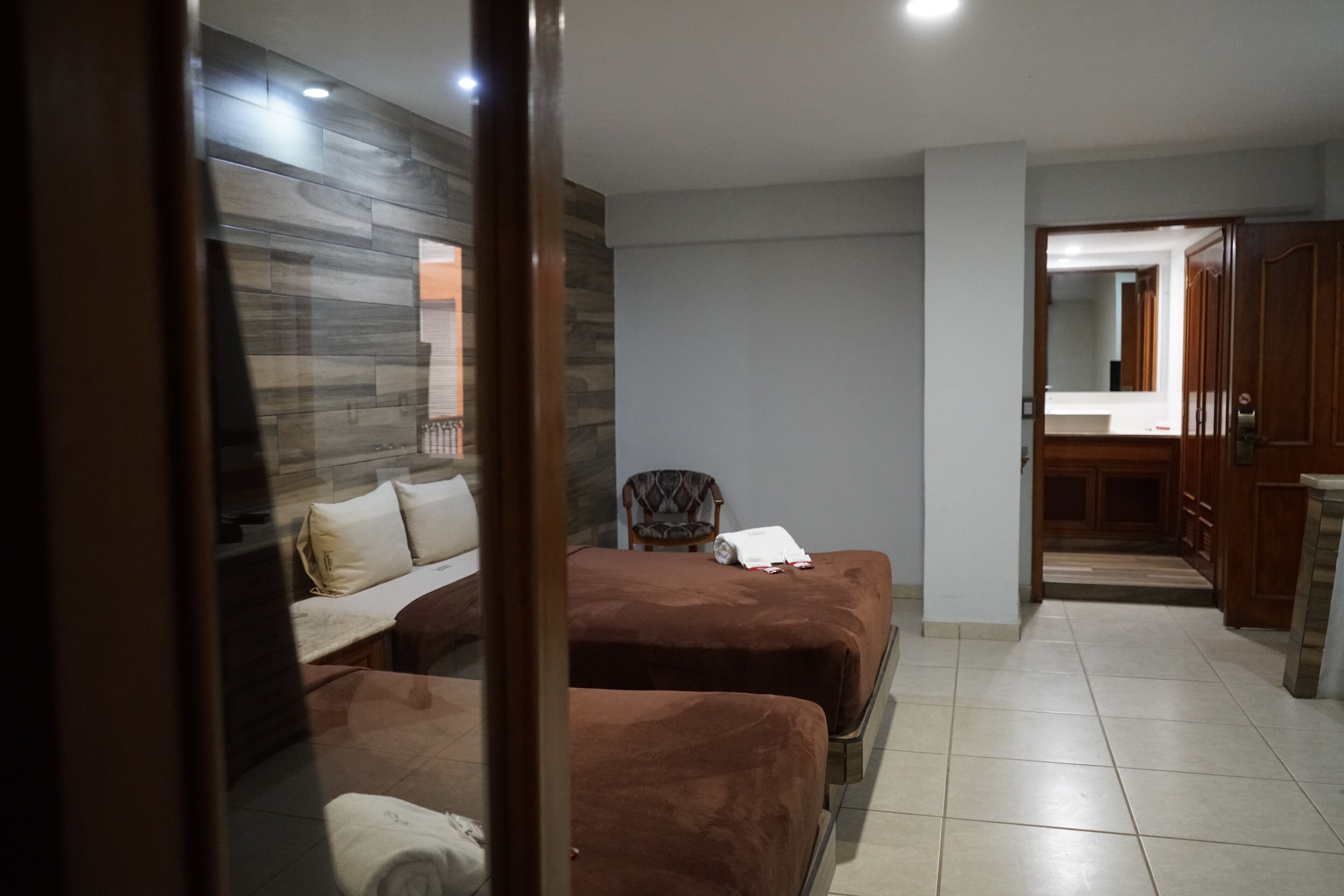 A hotel room with two beds, a chair, a wooden wardrobe, and a bathroom visible through a glass door.