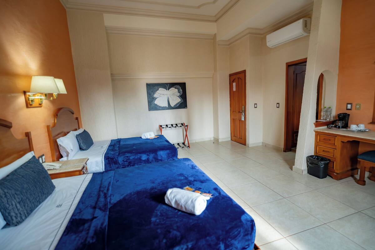 A hotel room with two beds, a desk, a chair, a lamp, a painting, and a trash bin.