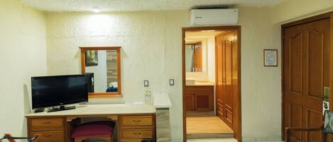 Una habitación con un escritorio, televisor, basurero, silla y puerta de madera.
