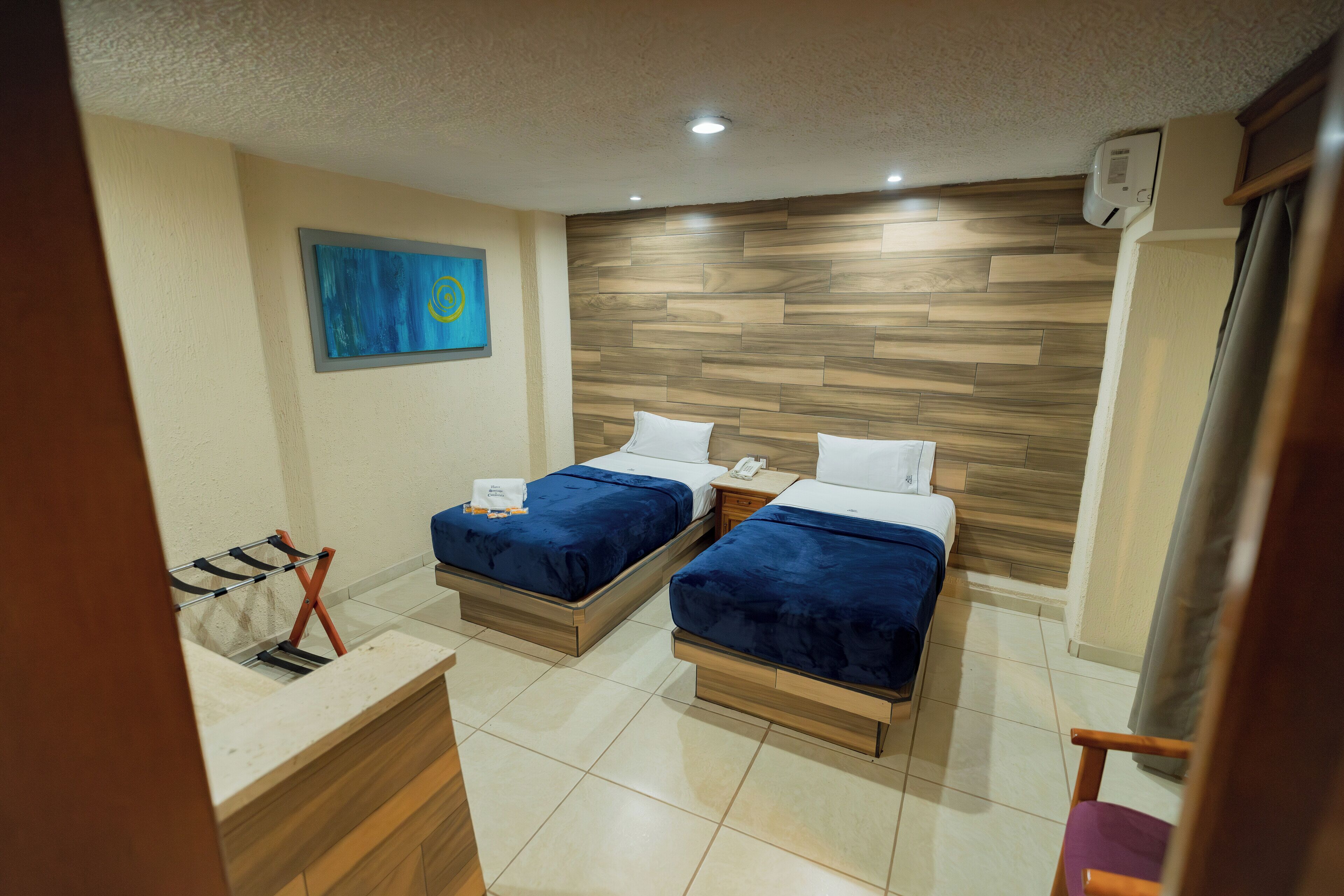 Ein Hotelzimmer mit zwei Betten, einem Holzkopfstück, einem blauen Gemälde, einem Waschbecken und einem Stuhl.