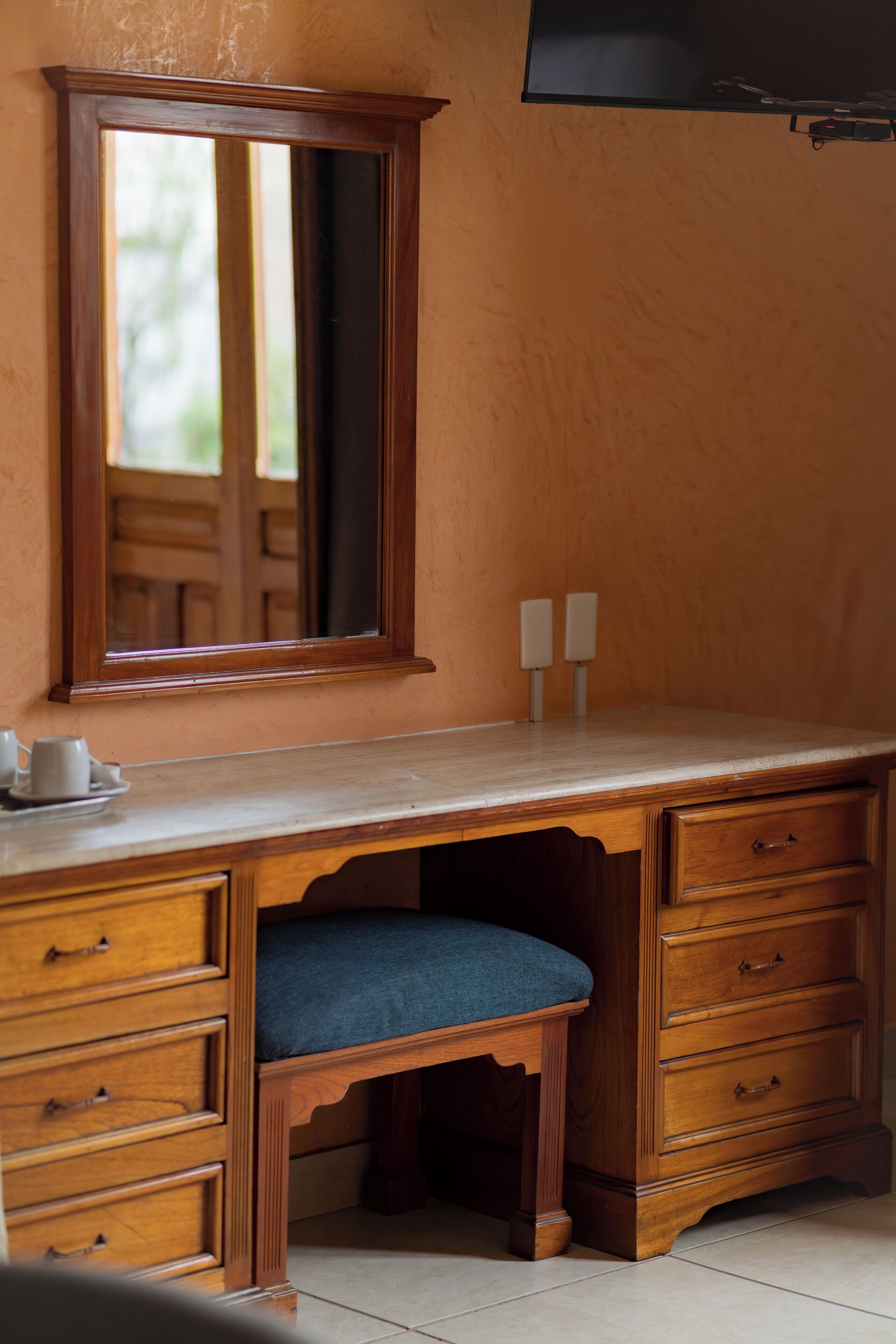 Un escritorio de madera con cajones y un espejo, un asiento acolchado azul y dos velas fijadas a la pared.