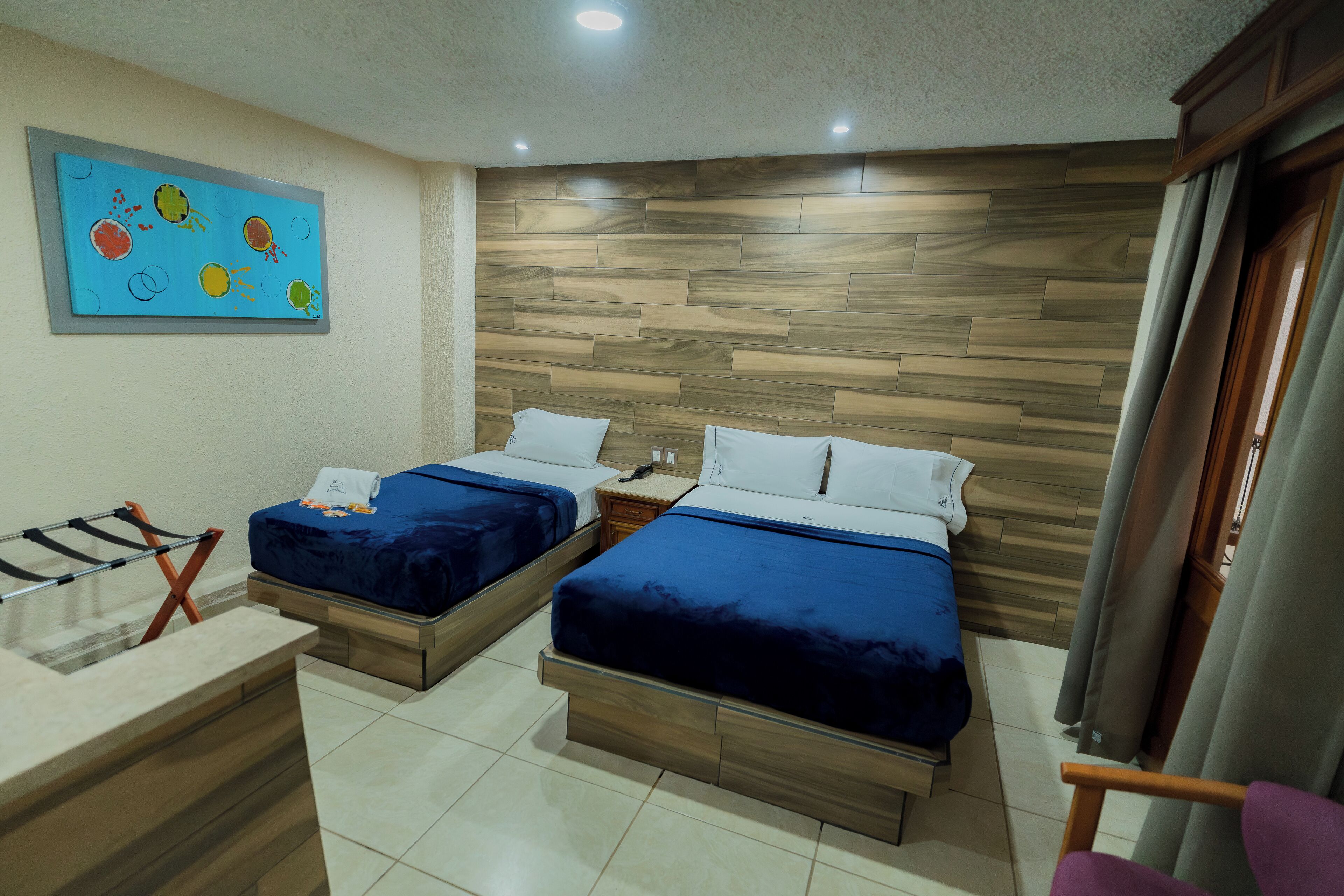 Habitación de hotel con dos camas, cabecero de madera, un escritorio pequeño y un cuadro de colores en la pared.