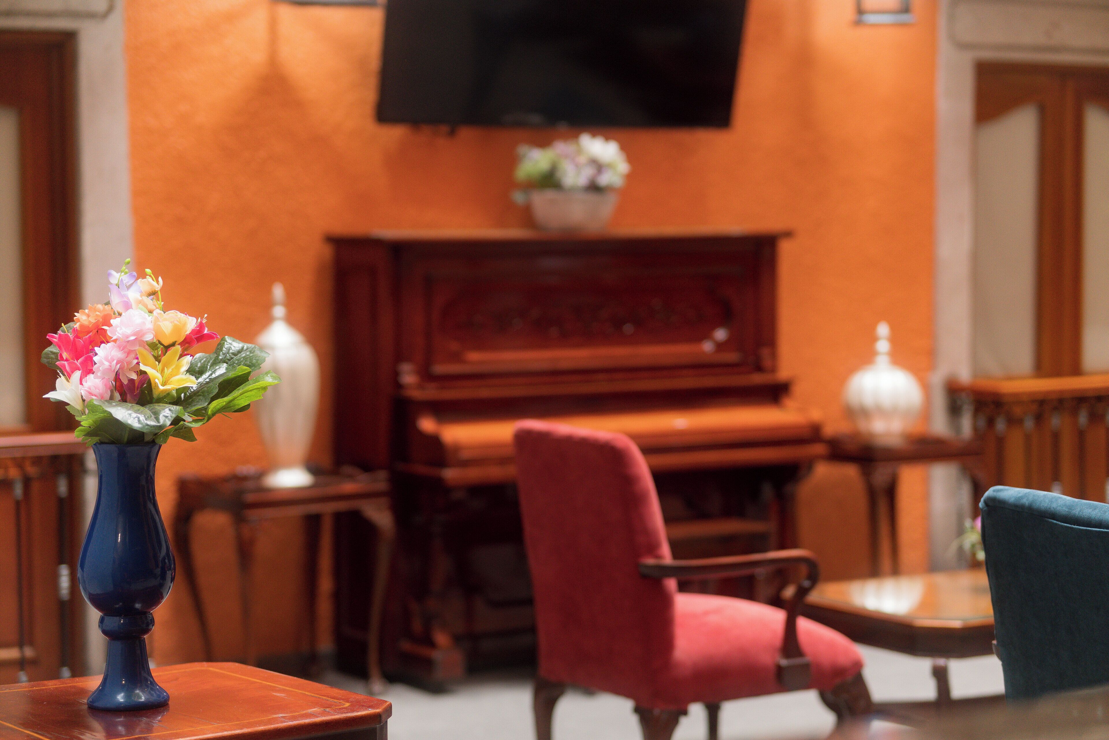 Una habitación con un piano de madera, una silla roja, un florero azul con flores y un televisor montado en la pared.
