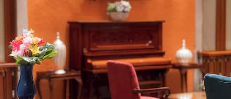 A room with a wooden piano, a red chair, a blue vase with flowers, and a television mounted on the wall.