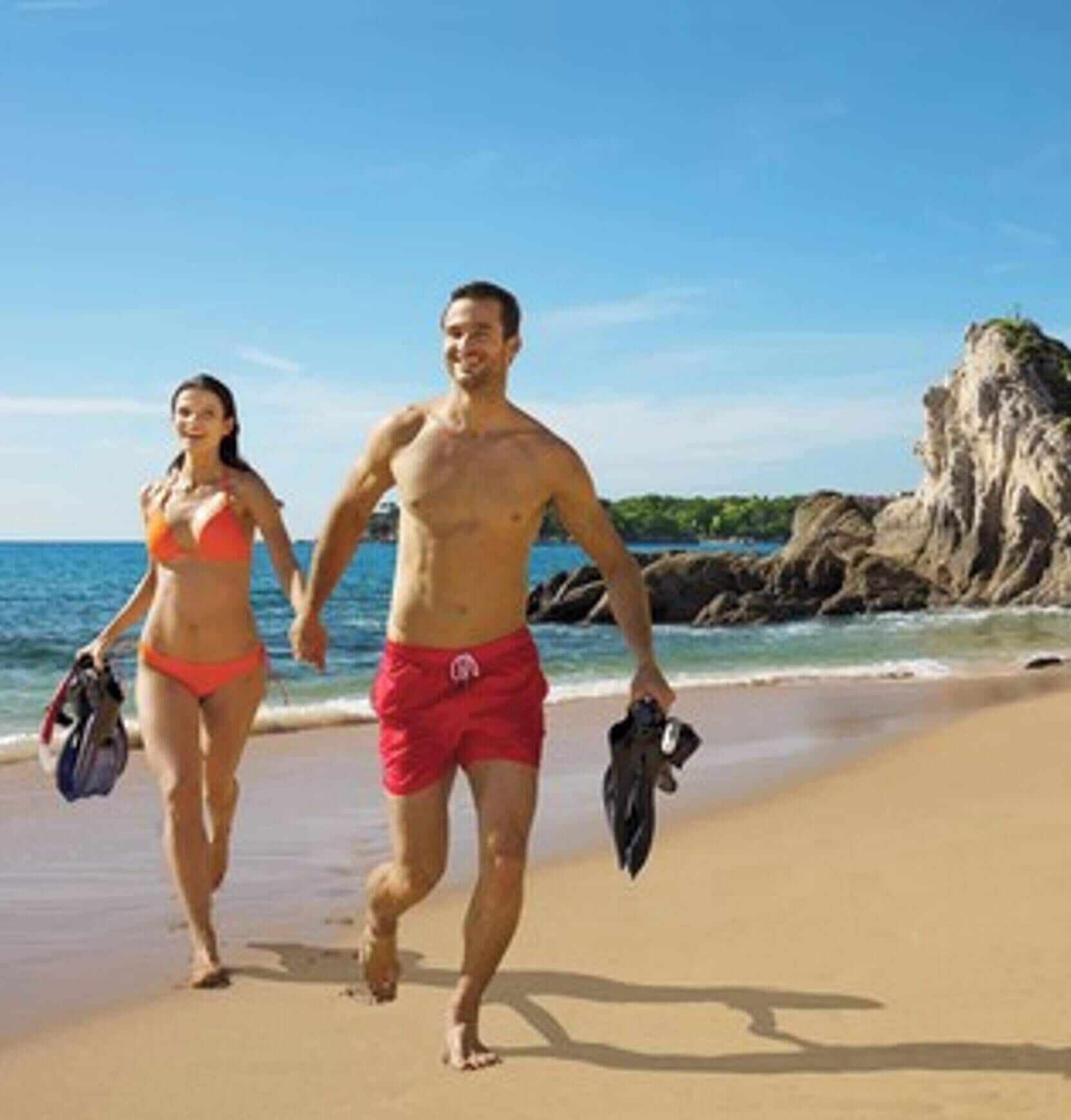 Two people walking on a sandy beach with a rocky cliff in the background.