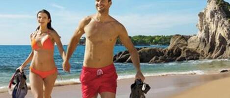 Two people walking on a sandy beach with a rocky cliff in the background.