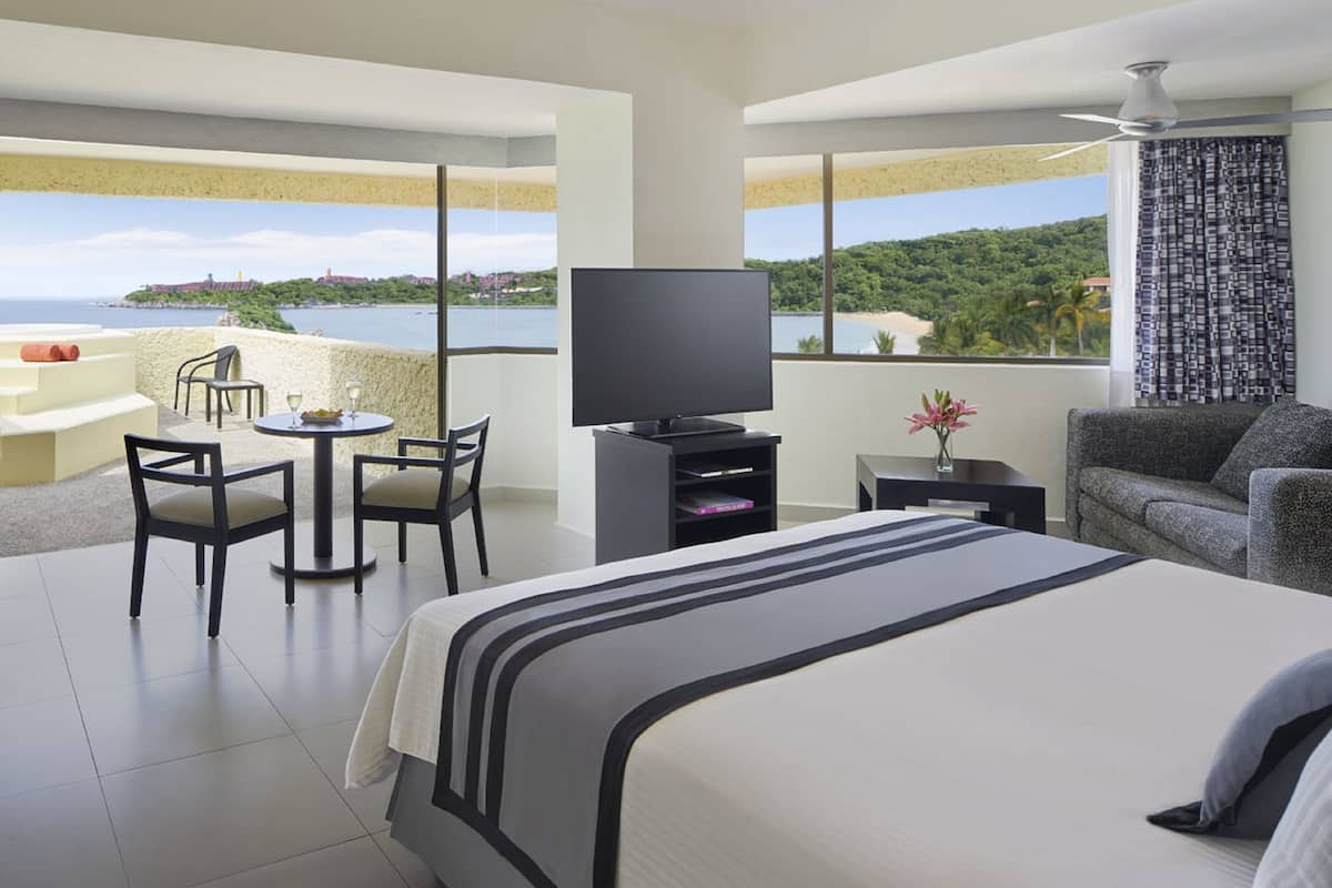 Ein Hotelzimmer mit einem großen Bett, einem Fernseher, einem Balkon mit Meerblick und einem Essbereich mit Stühlen und einem Tisch.