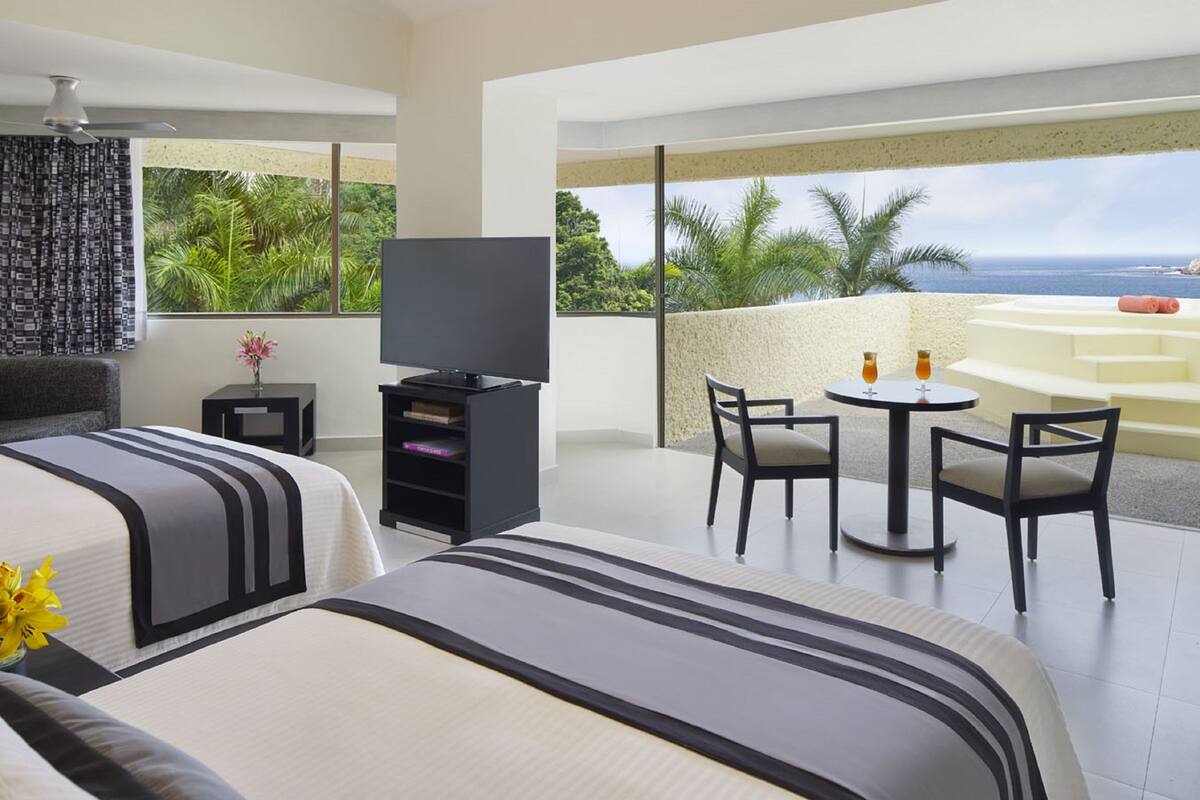 Ein Hotelzimmer mit zwei Betten, einem Fernseher, einem kleinen Tisch mit Stühlen und einem Balkon mit Blick auf den Ozean und Palmen.