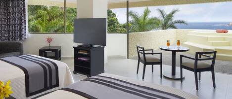 A hotel room with two beds, a TV, a small table with chairs, and a balcony with a view of the ocean and palm trees.