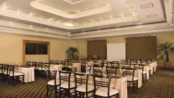 A conference room with multiple tables and chairs, a projector screen, and a large potted plant.