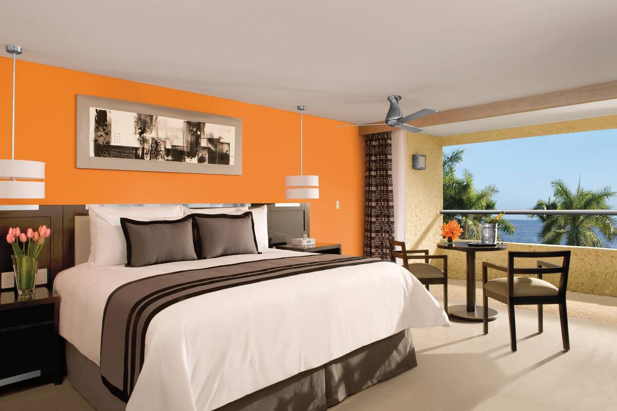 Ein Hotelzimmer mit einem großen Bett, einem Schreibtisch mit Stuhl, einem Balkon mit Tisch und Stühlen sowie Blick aufs Meer.