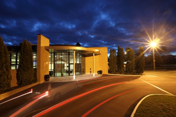 A modern building with a glass facade, a brick exterior, and a parking lot with light trails from moving vehicles. - Distinction Rotorua Hotel and Conference Centre (Rotorua)