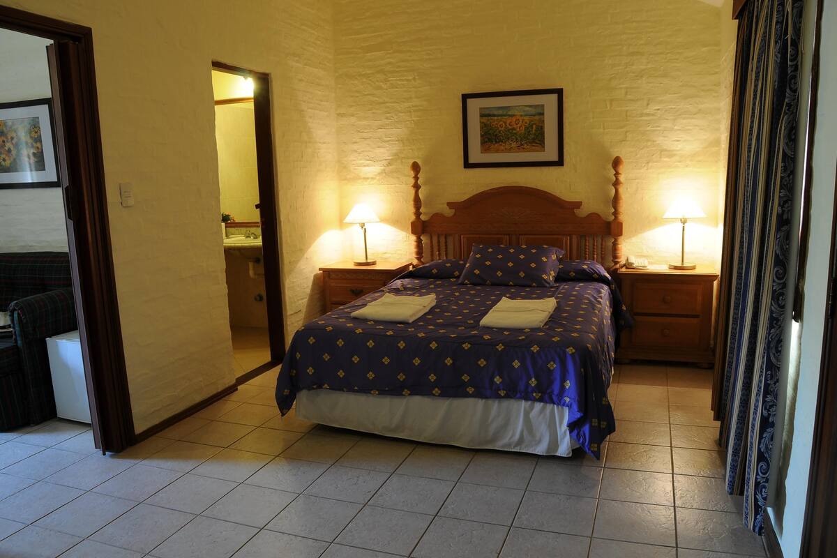 A bedroom with a bed, two bedside tables with lamps, a framed picture on the wall, and a bathroom visible through an open door.