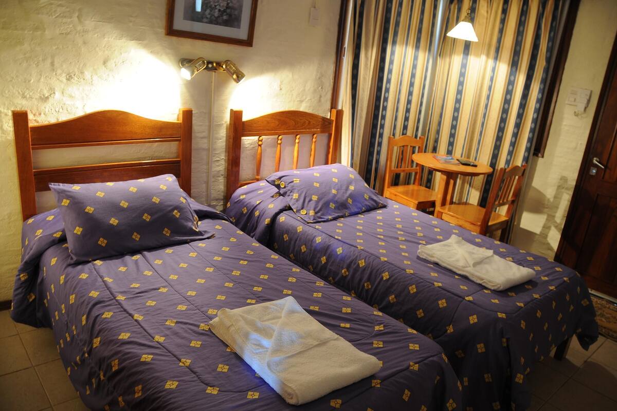 A room with two wooden beds with purple bedding, a small wooden table, and a chair.