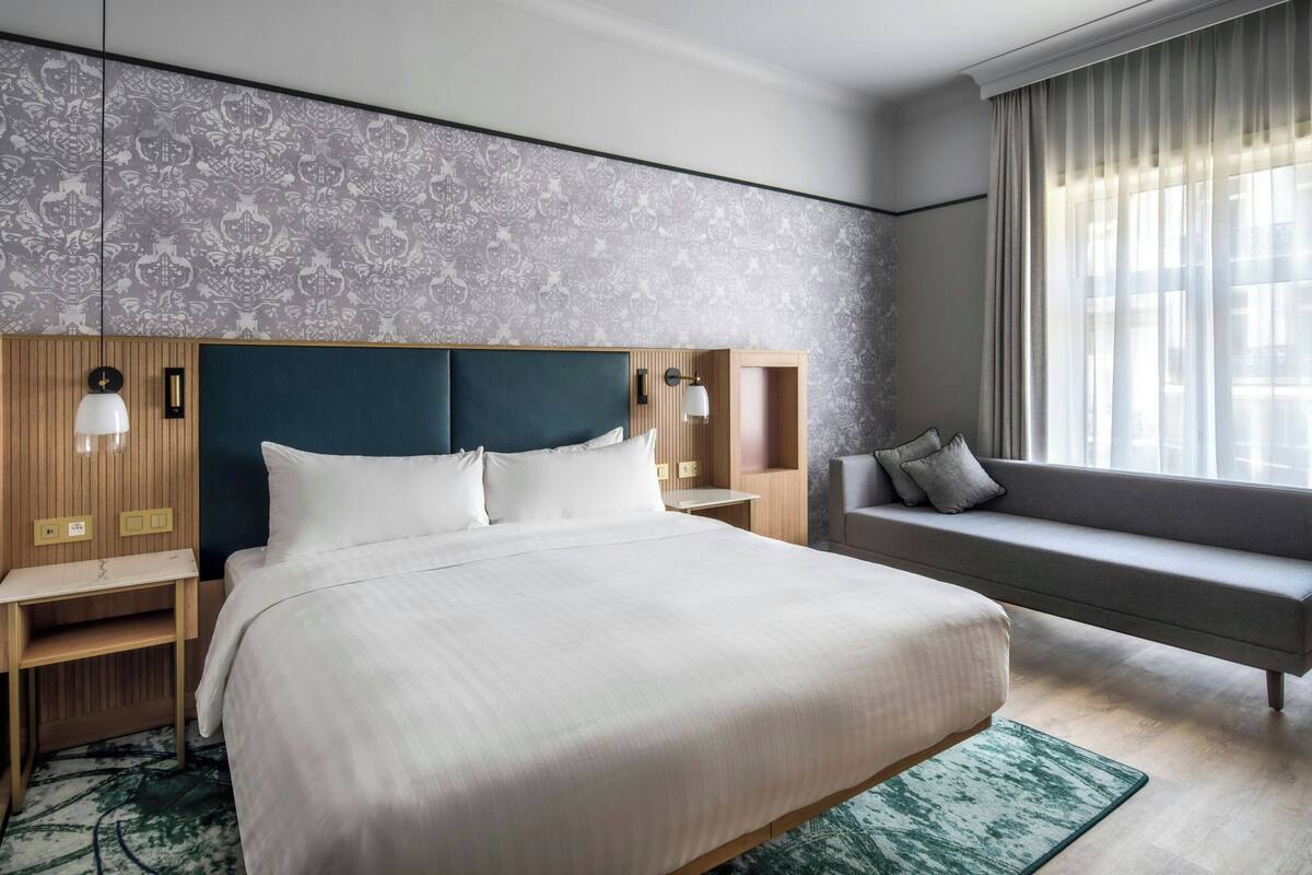 A hotel room with a large bed, bedside tables, a sofa, a window with curtains, and a patterned wall.