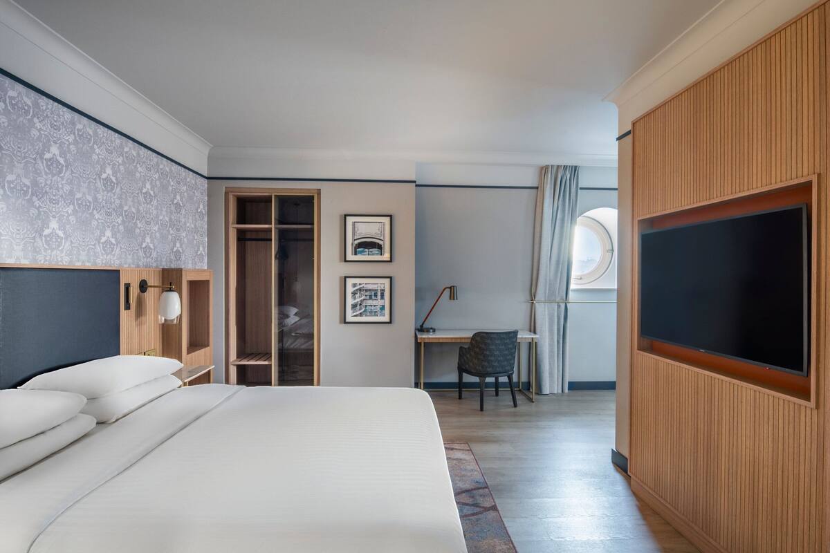 A modern hotel room with a large bed, a desk with a chair, a wardrobe, and a television mounted on the wall.