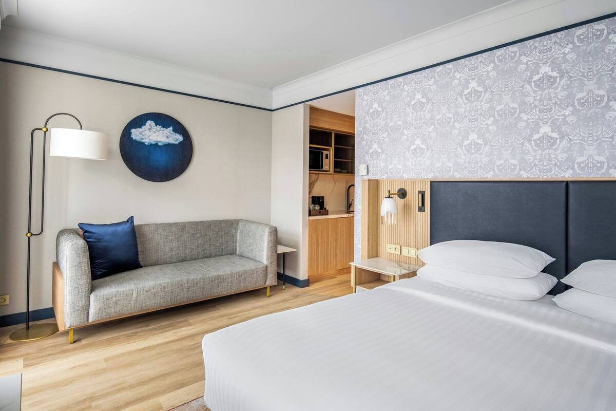 A hotel room with a bed, a sofa, a desk, a lamp, a wall-mounted artwork, and a kitchen area.