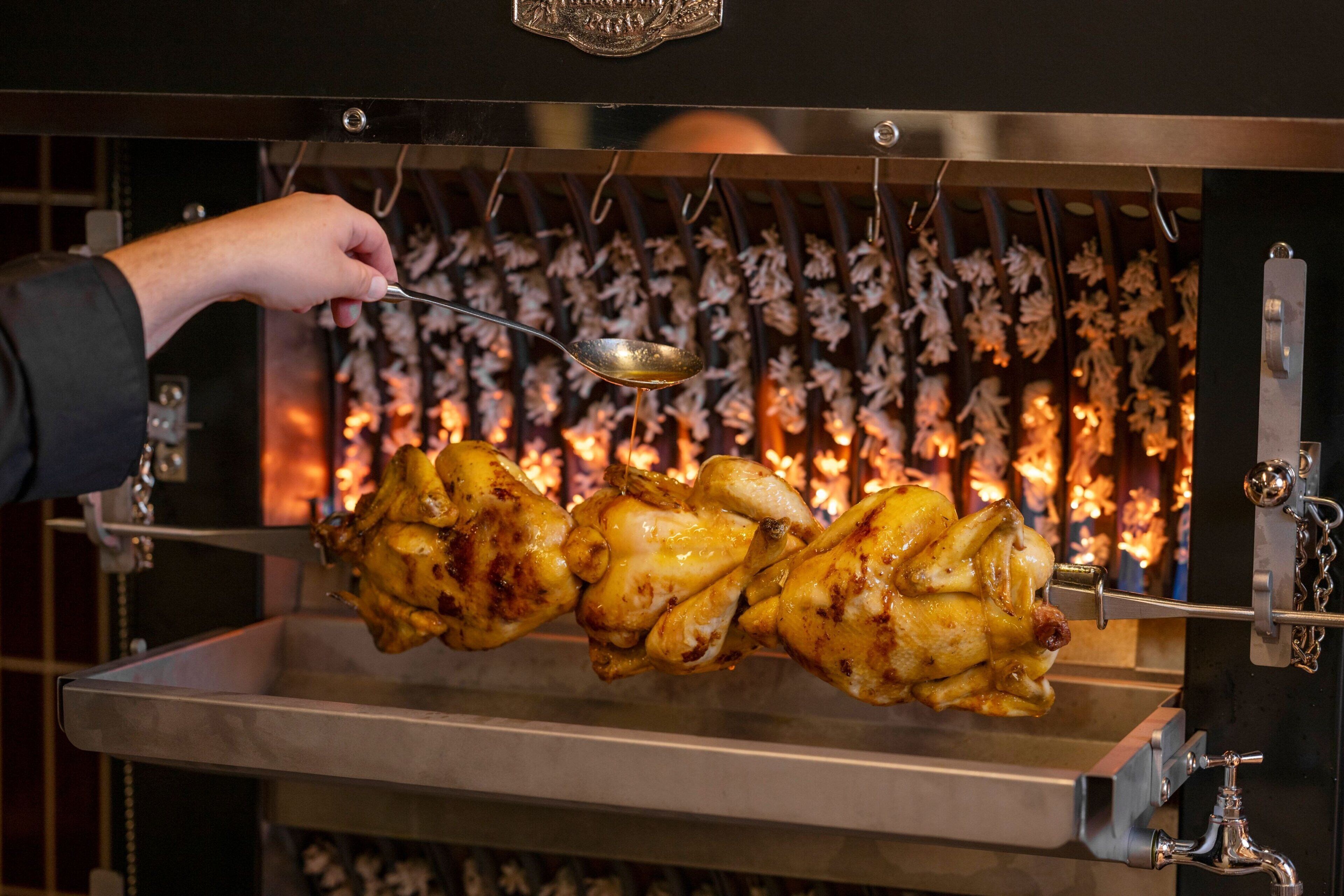 A person using a spoon to handle a large, roasted chicken on a spit in a commercial kitchen.