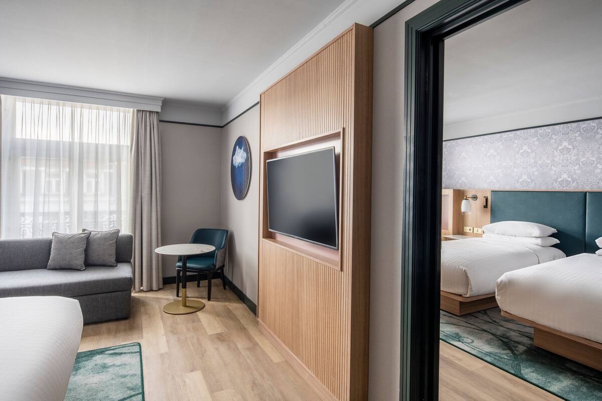 A hotel room with a sofa, a round table, a TV mounted on a wooden panel, and two beds with headboards.