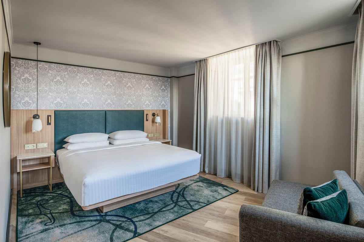 A hotel room with a bed, a nightstand, a chair, a patterned wall, and a window with curtains.