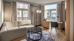 Una habitación de hotel moderna con sofá, una pequeña mesa redonda, un escritorio y una silla. Hay ventanas amplias con cortinas, una lámpara de pie y vista a la ciudad.