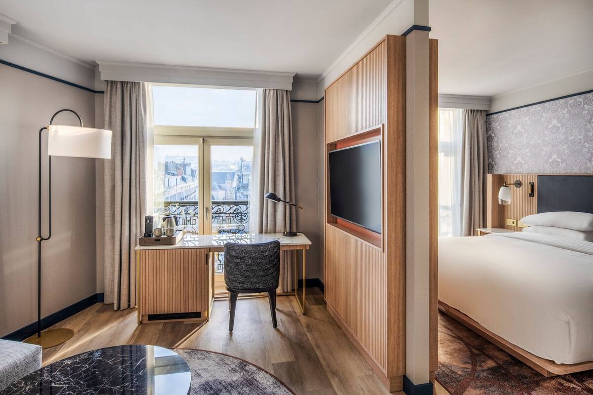 A hotel room with a bed, desk, chair, and a large window with a city view.