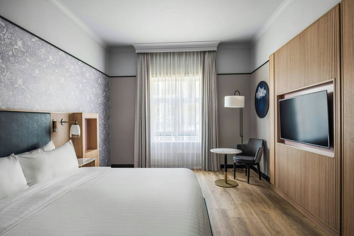 A hotel room with a large bed, a TV mounted on the wall, a small table with two chairs, and a window with sheer curtains.