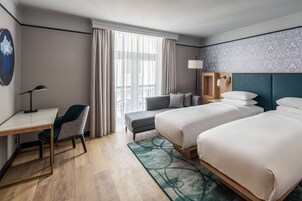 Et hotelværelse med to senge, et skrivebord, en stol og et stort vindue med gardiner.