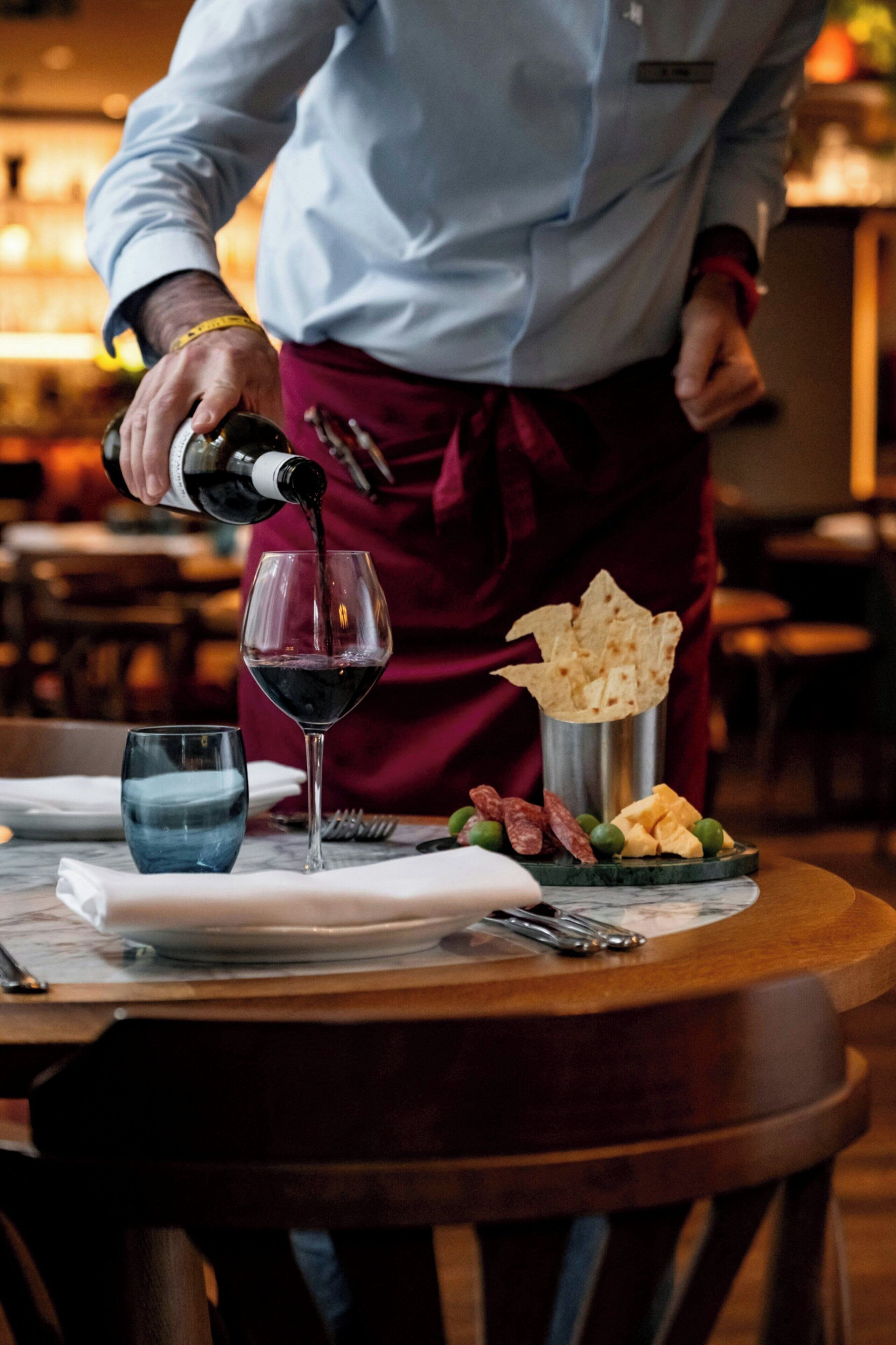 A person pouring wine into a glass on a table with plates and a metal cup.