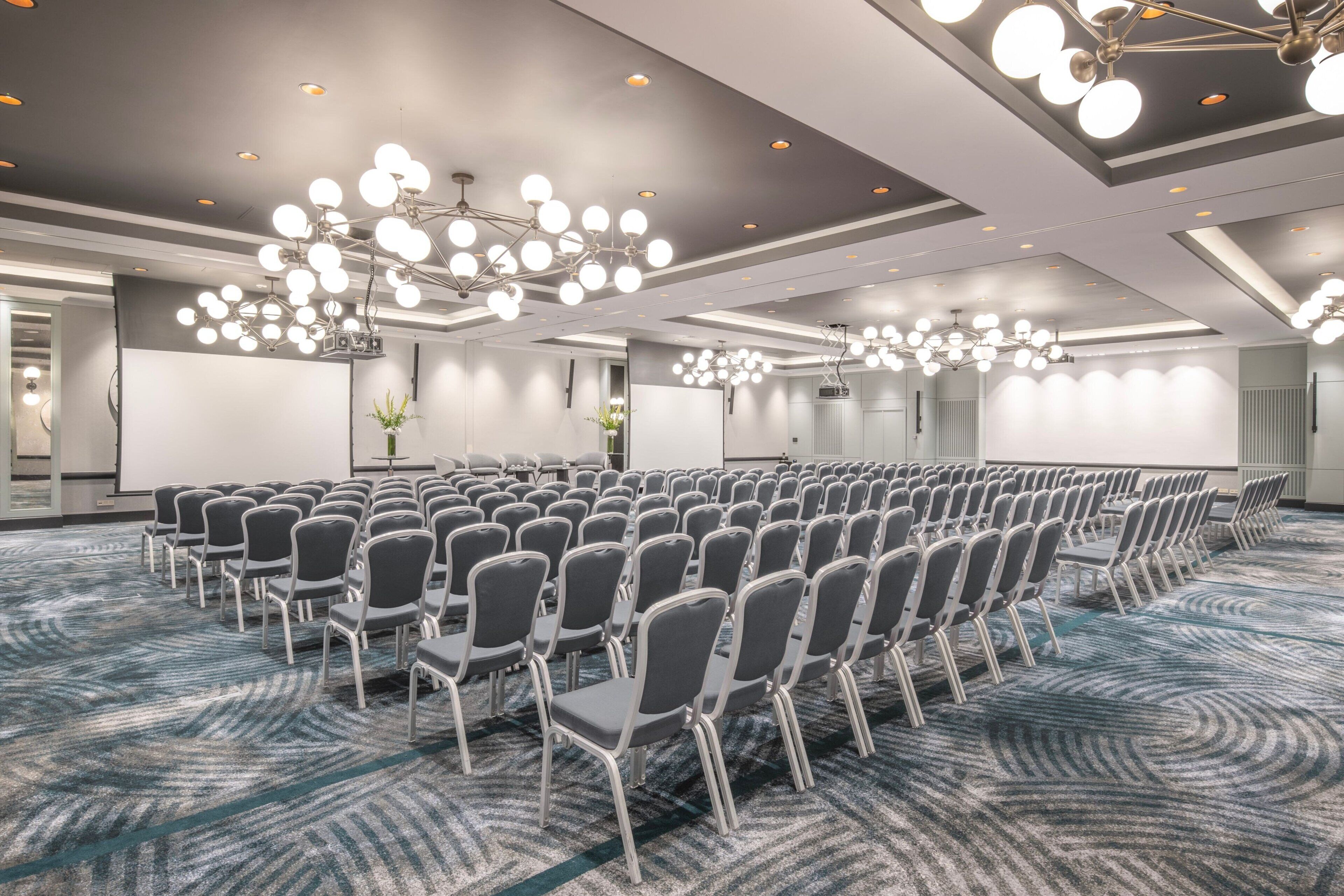Un salón de conferencias con filas de sillas, luces en el techo y piso alfombrado.