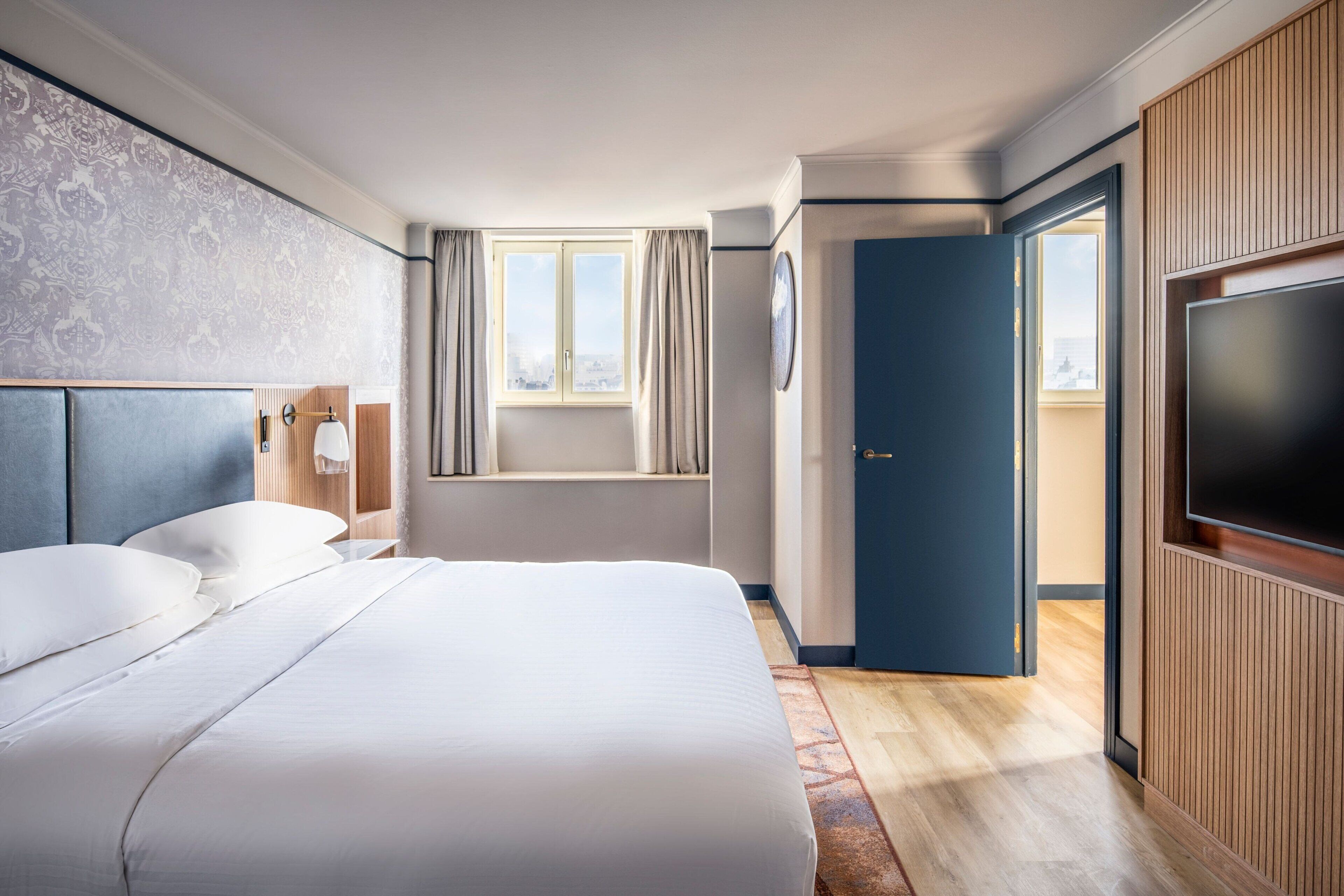 A hotel room with a large bed, a television, a window with curtains, and a door leading to another room.