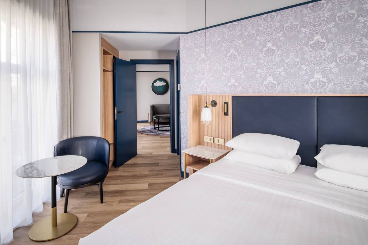 A hotel room with a bed, a small table, a chair, and a desk. There is a door leading to another room.