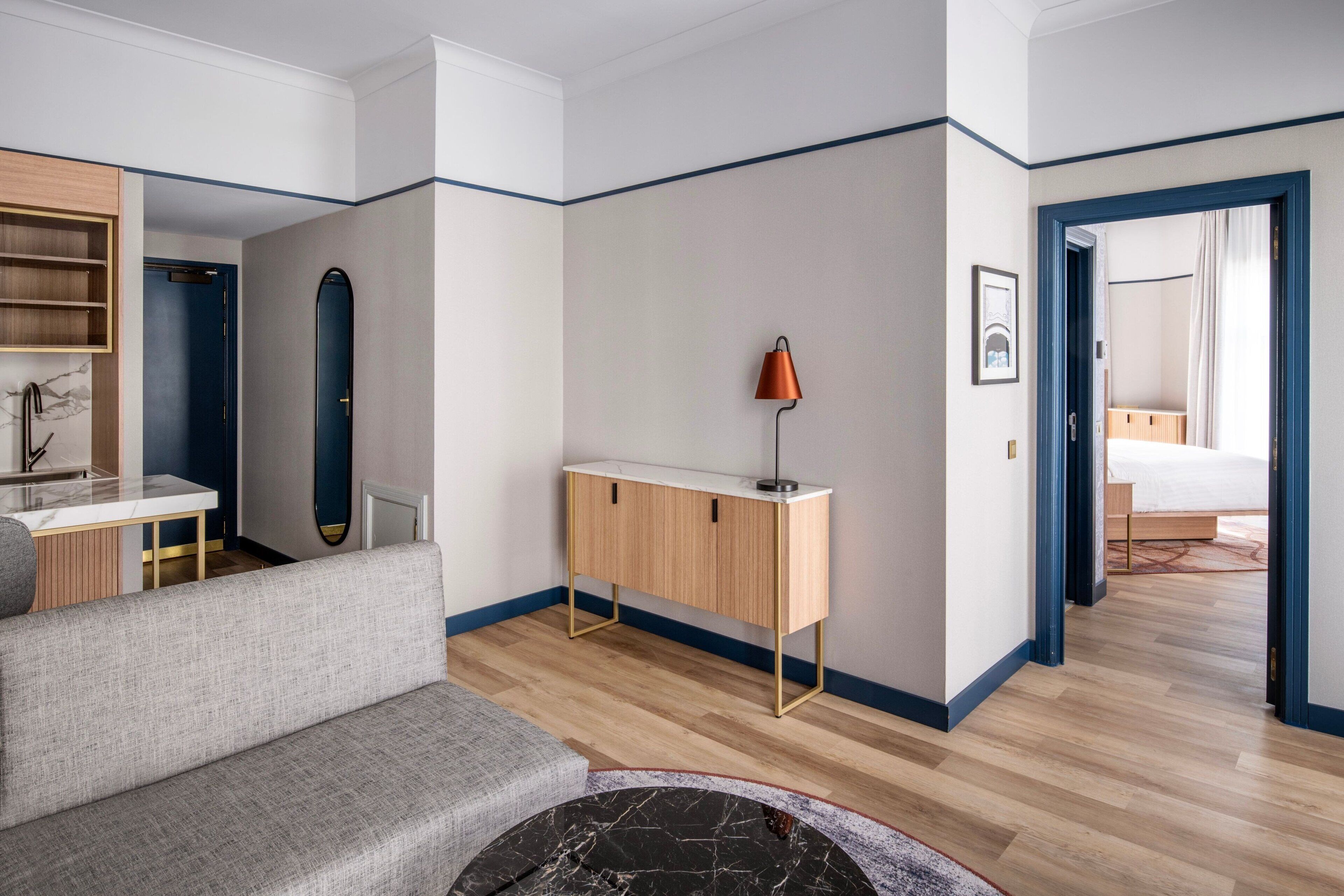 A modern hotel room with a grey sofa, a wooden desk, a mirror, and a bed in the background.