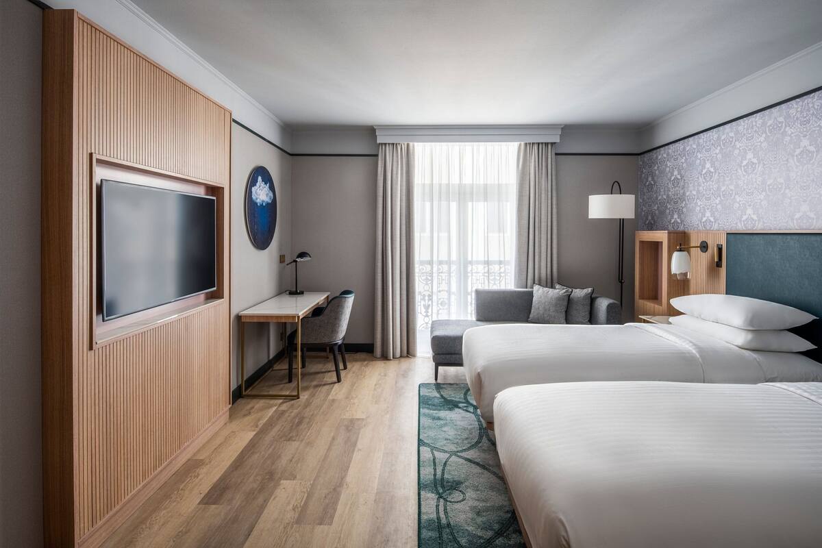 A hotel room with two beds, a desk, a TV, and a view of the city.
